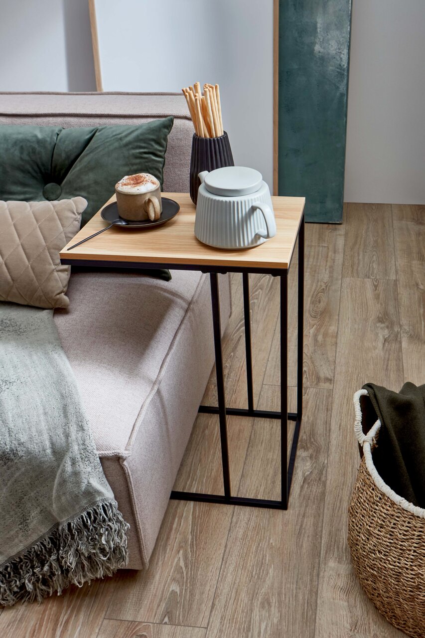 MID.YOU Couchtisch SMALL Schmaler Beistelltisch mit heller Holzplatte und schwarzem Metallgestell neben einem Sofa, darauf Tasse und Keramikdose, fotografiert in seitlicher Perspektive auf das Produkt.