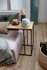 Schmaler Beistelltisch mit heller Holzplatte und schwarzem Metallgestell neben einem Sofa, darauf Tasse und Keramikdose, fotografiert in seitlicher Perspektive auf das Produkt.