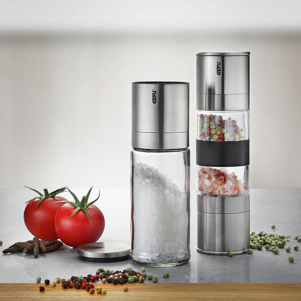 Geschenkset LAMOLA mit Salz- und Pfeffermühle aus Edelstahl, gefüllt mit grobem Salz und buntem Pfeffer, auf einer Küchenarbeitsplatte mit Tomaten und Pfefferkörnern im Vordergrund, seitliche Perspektive.