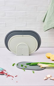 Drei Schneidbretter in verschiedenen Größen und Farben, aufrecht stehend vor einer weißen Kachelwand. Im Vordergrund liegt ein Schneidbrett mit Frühlingszwiebeln, daneben Pfefferkörner, Zitronenhälften und Knoblauch. Perspektive: Frontalansicht.