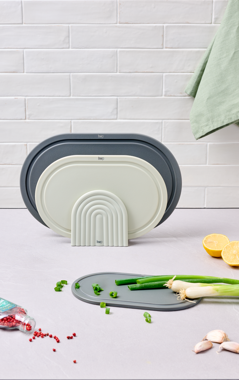 Homeware Schneidbrettset Drei Schneidbretter in verschiedenen Größen und Farben, aufrecht stehend vor einer weißen Kachelwand. Im Vordergrund liegt ein Schneidbrett mit Frühlingszwiebeln, daneben Pfefferkörner, Zitronenhälften und Knoblauch. Perspektive: Frontalansicht.