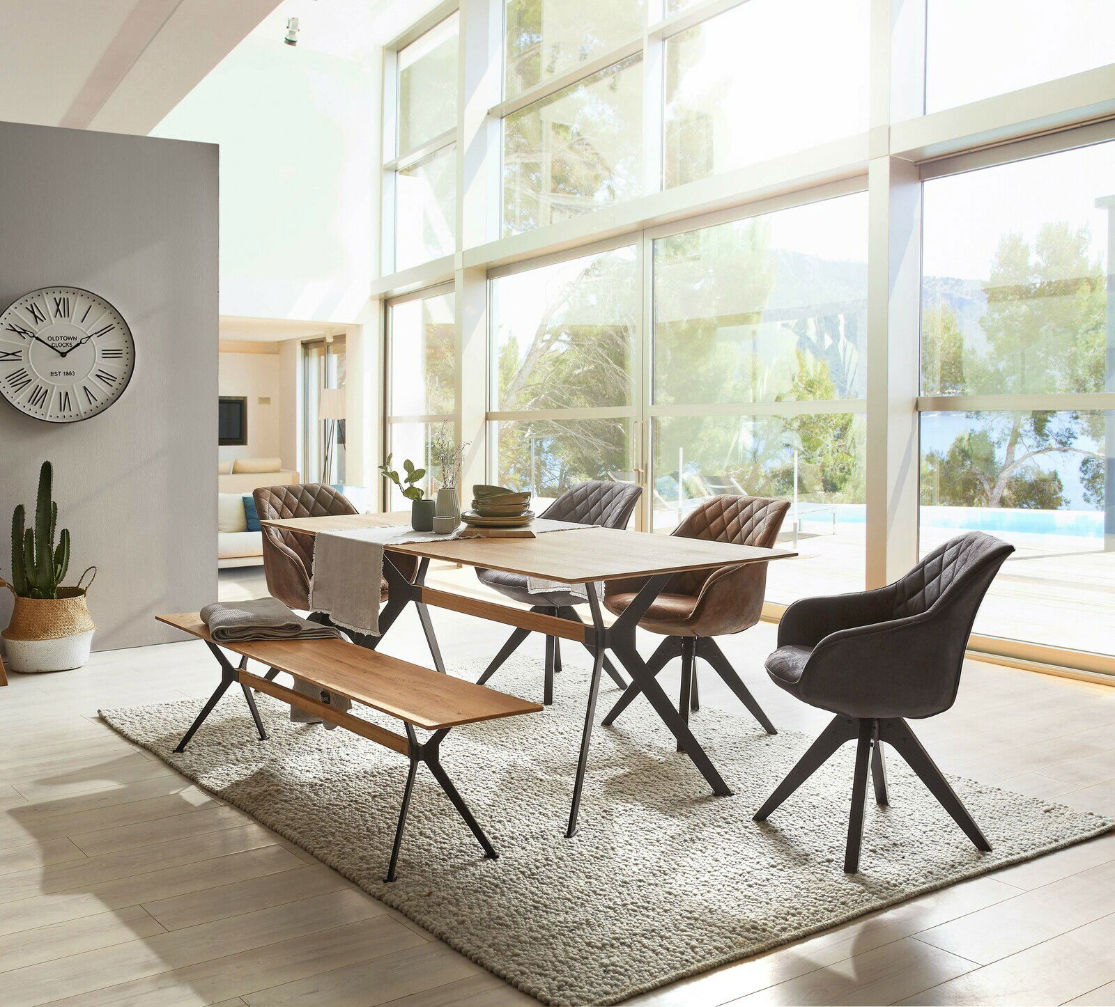 Moderner Esstisch aus Holz mit Metallbeinen, umgeben von stilvollen Stühlen und einer Bank, in einem hellen Raum mit großen Fenstern und Blick auf die Natur. Perspektive von der Seite.
