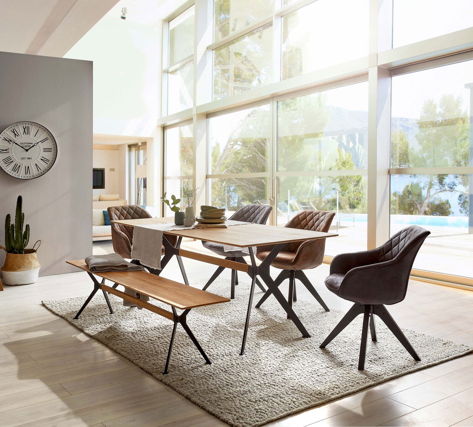 Linea Natura Esstisch 120x80 DÜSSELDORF Moderner Esstisch aus Holz mit Metallbeinen, umgeben von stilvollen Stühlen und einer Bank, in einem hellen Raum mit großen Fenstern und Blick auf die Natur. Perspektive von der Seite.