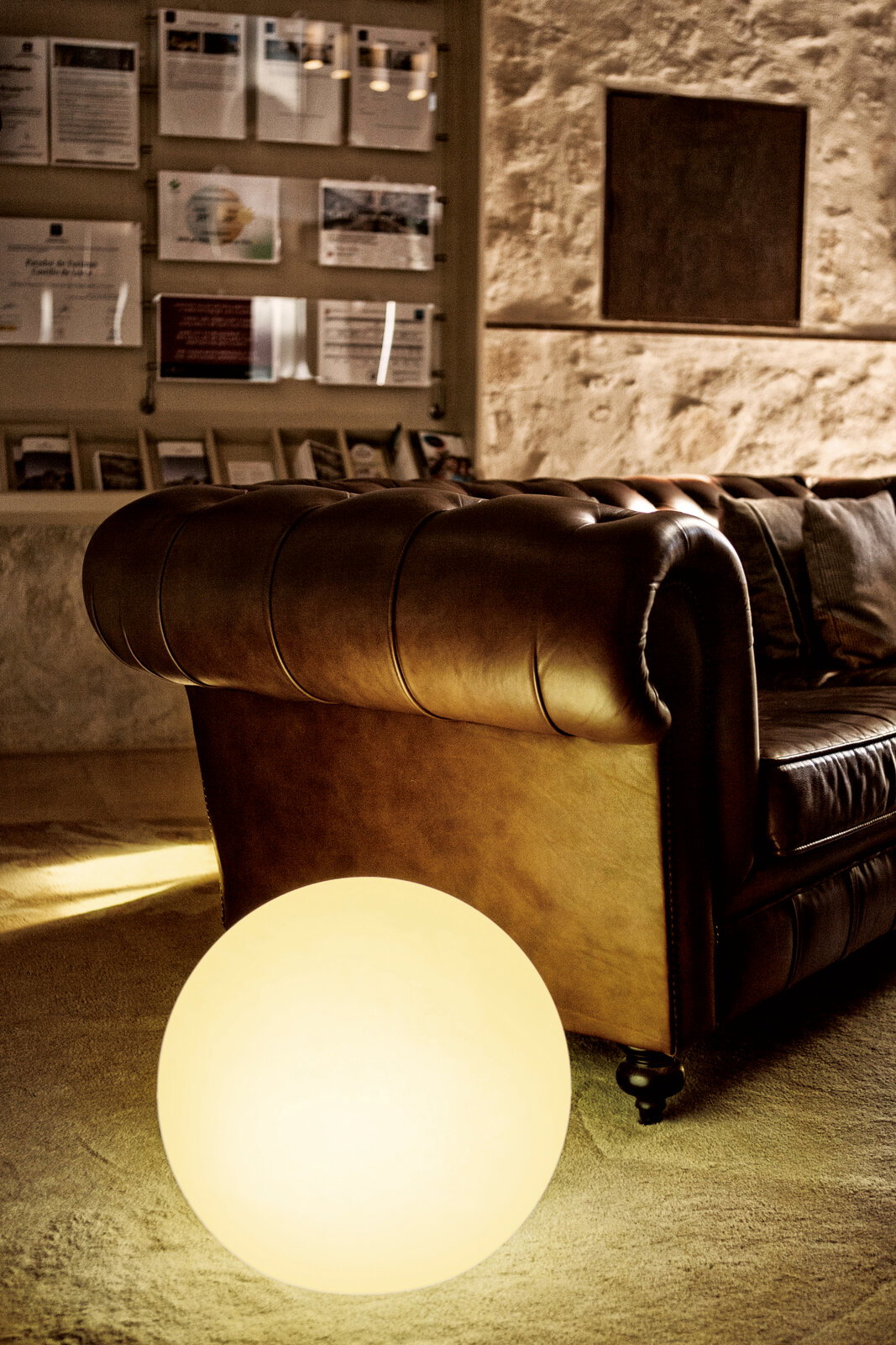 Braunes Chesterfield-Ledersofa in gemütlicher Lounge, im Vordergrund leuchtende Kugellampe auf dem Boden; Perspektive: seitliche Nahaufnahme auf Armlehne und Sitzfläche