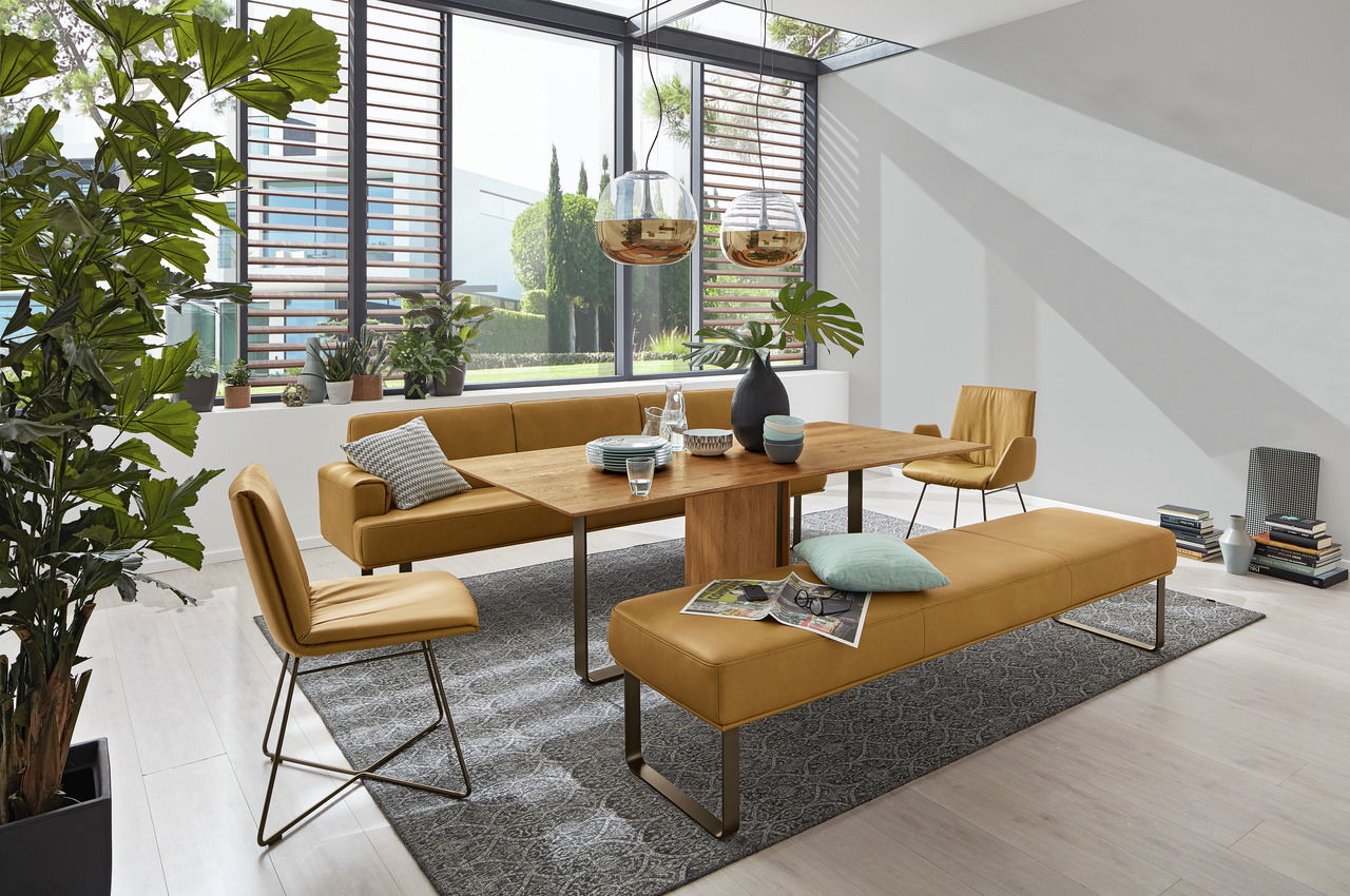 Moderne Esszimmer-Szene mit einem Holztisch, umgeben von senffarbenen Stühlen und Bänken, mit Blick auf große Fenster und viel Tageslicht.