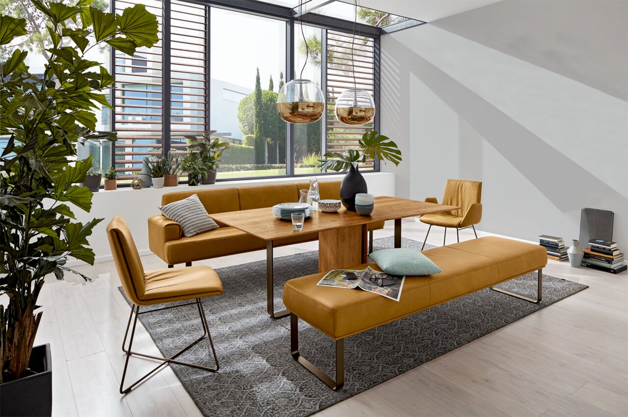 Moderne Esszimmer-Szene mit einem Holztisch, umgeben von senffarbenen Stühlen und Bänken, mit Blick auf große Fenster und viel Tageslicht.