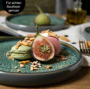 Nahaufnahme eines grünen Tellers mit einer Feige, einem grünen und einem rosa Macaron, dekoriert mit Nüssen und getrockneten Früchten. Im Hintergrund unscharf ein weiterer Teller und eine Flasche Öl. Perspektive von schräg oben.