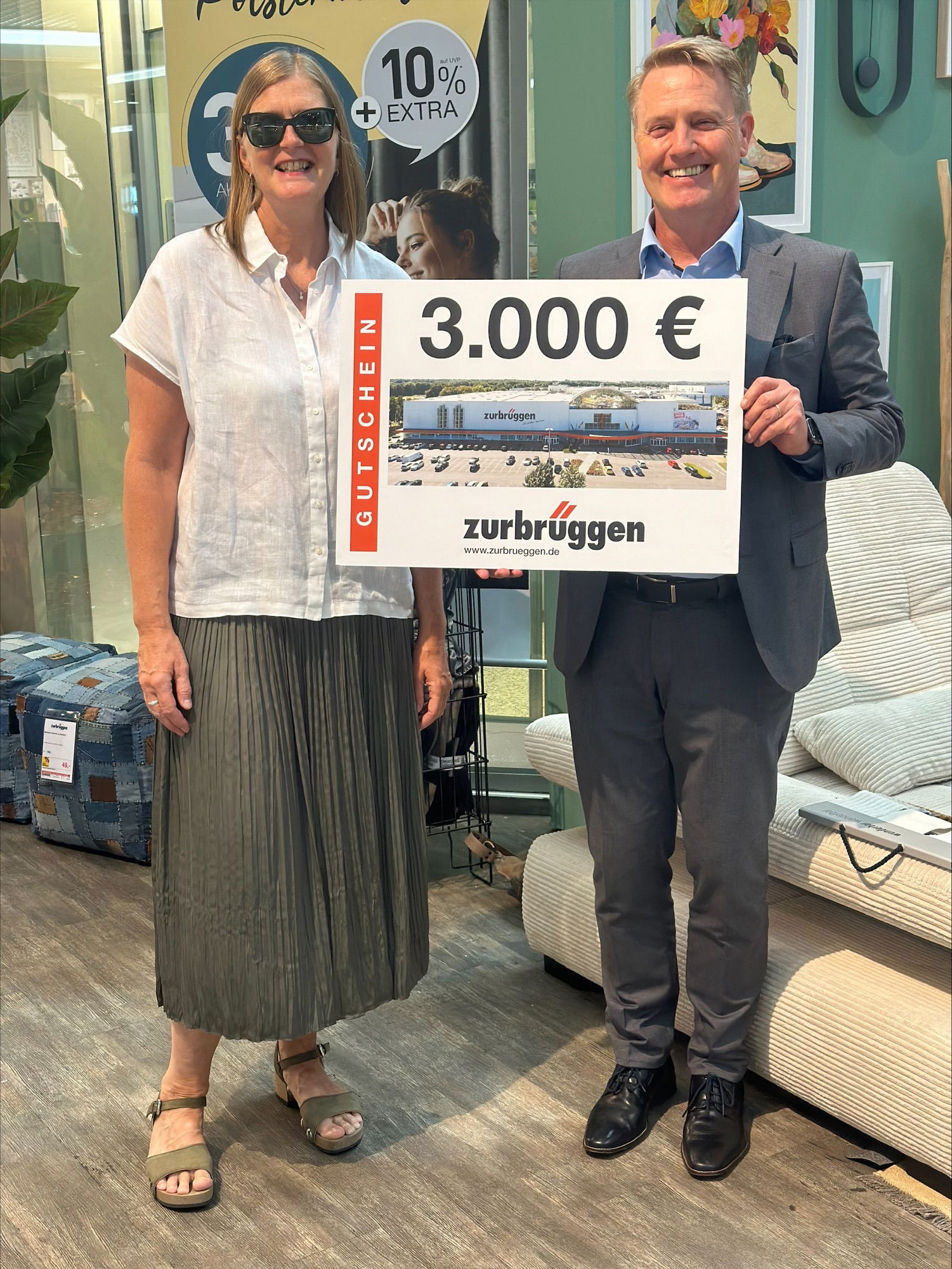 Das Bild zeigt zwei Personen, die einen großen Gutschein in der Hand halten. Auf dem Gutschein steht „3.000 €“ und das Logo von „Zurbrüggen“. Es scheint, dass eine Spende oder ein Gewinn übergeben wurde.