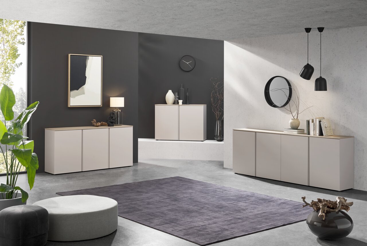 Voleo XL-dressoir MILTON Moderne woonkamerinrichting met drie lichtgrijze dressoirs met houten bovenblad, decoratie en wandspiegels, gefotografeerd in een schuin vooraanzicht vanuit een iets hoger standpunt.
