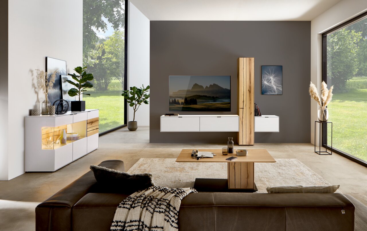 Moderne Wohnzimmer-Wandkombination mit hängenden weißen Schränken und einem vertikalen Holzelement, frontal betrachtet. Der Raum ist hell und minimalistisch eingerichtet mit großen Fenstern, die einen Blick ins Grüne bieten.