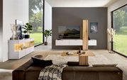 Moderne Wohnzimmer-Wandkombination mit hängenden weißen Schränken und einem vertikalen Holzelement, frontal betrachtet. Der Raum ist hell und minimalistisch eingerichtet mit großen Fenstern, die einen Blick ins Grüne bieten.