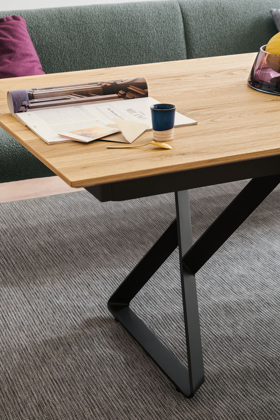 Nahaufnahme eines modernen Esstisches mit Holzplatte und schwarzem Metallgestell, dekoriert mit Zeitschriften, einer Tasse und einem goldenen Löffel, aus seitlicher Perspektive.