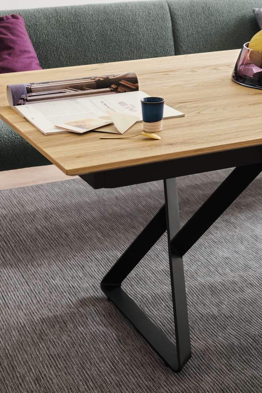 Nahaufnahme eines modernen Esstisches mit Holzplatte und schwarzem Metallgestell, dekoriert mit Zeitschriften, einer Tasse und einem goldenen Löffel, aus seitlicher Perspektive.