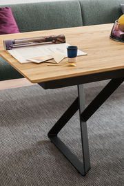 Nahaufnahme eines modernen Esstisches mit Holzplatte und schwarzem Metallgestell, dekoriert mit Zeitschriften, einer Tasse und einem goldenen Löffel, aus seitlicher Perspektive.