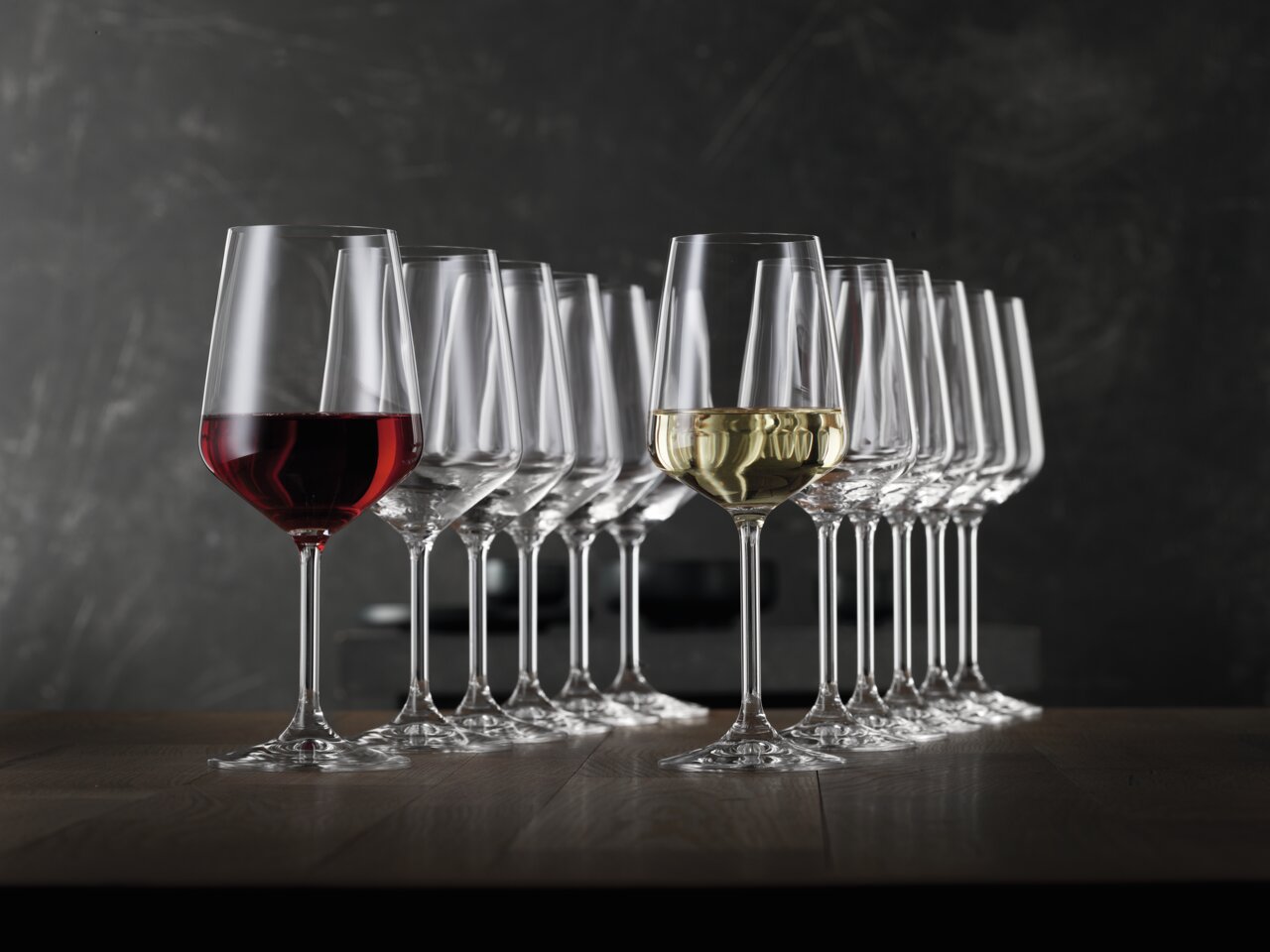 Eine Reihe von eleganten Kelchgläsern, teilweise mit Rot- und Weißwein gefüllt, auf einem Holztisch vor einem dunklen Hintergrund. Seitliche Perspektive.