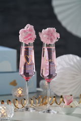 Zwei Sektgläser mit der Aufschrift 'Love' und einem Herzmotiv, gefüllt mit rosa Getränk und rosa Zuckerwatte, auf einem festlich dekorierten Tisch. Perspektive: Frontalansicht.