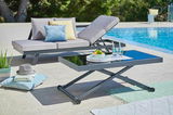 Moderner Gartentisch mit Glasplatte, im Vordergrund auf einer Terrasse neben einem Pool, umgeben von Kissen und einem Liegestuhl.