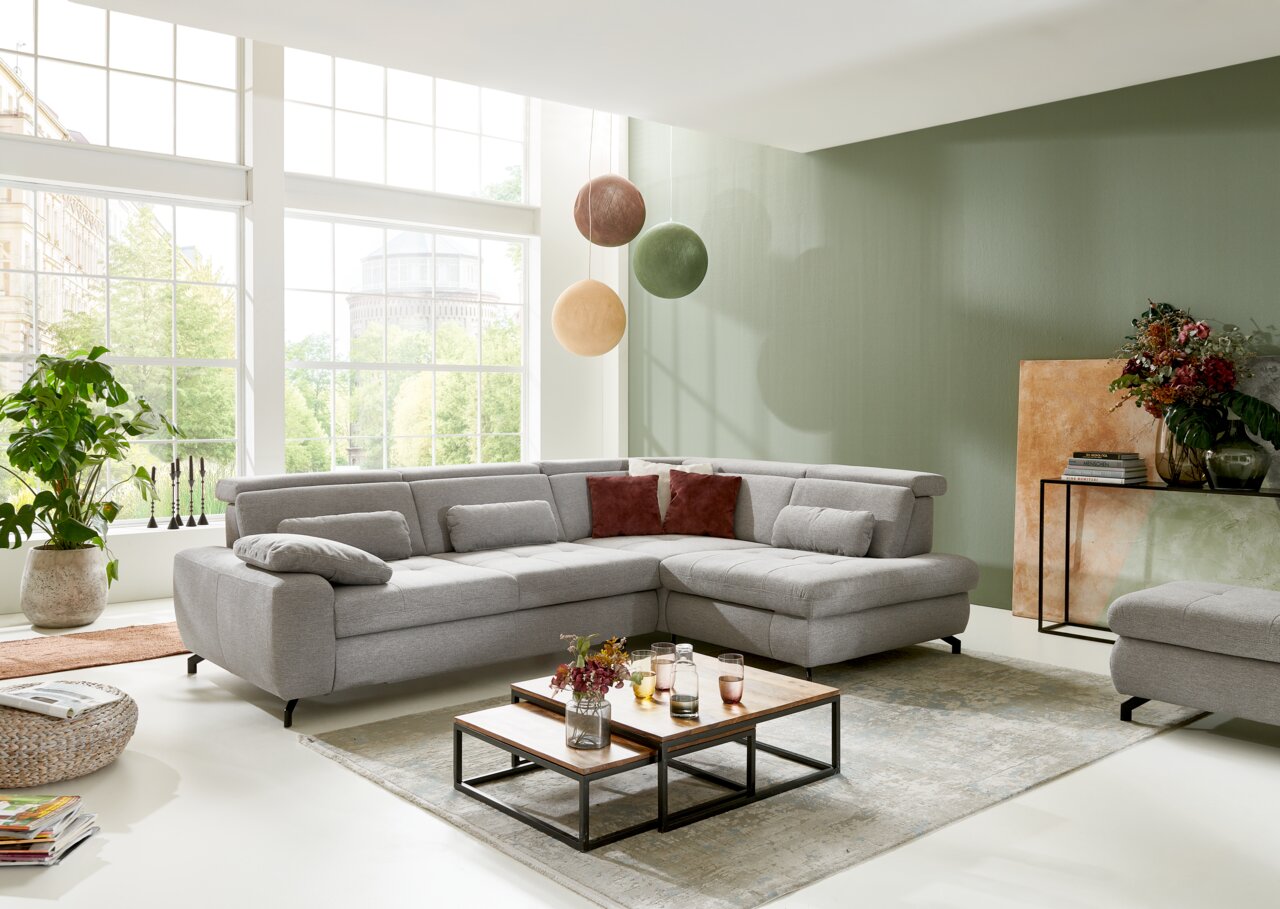 Graue Eckgarnitur in modernem Wohnzimmer mit großen Fenstern, von vorne betrachtet.