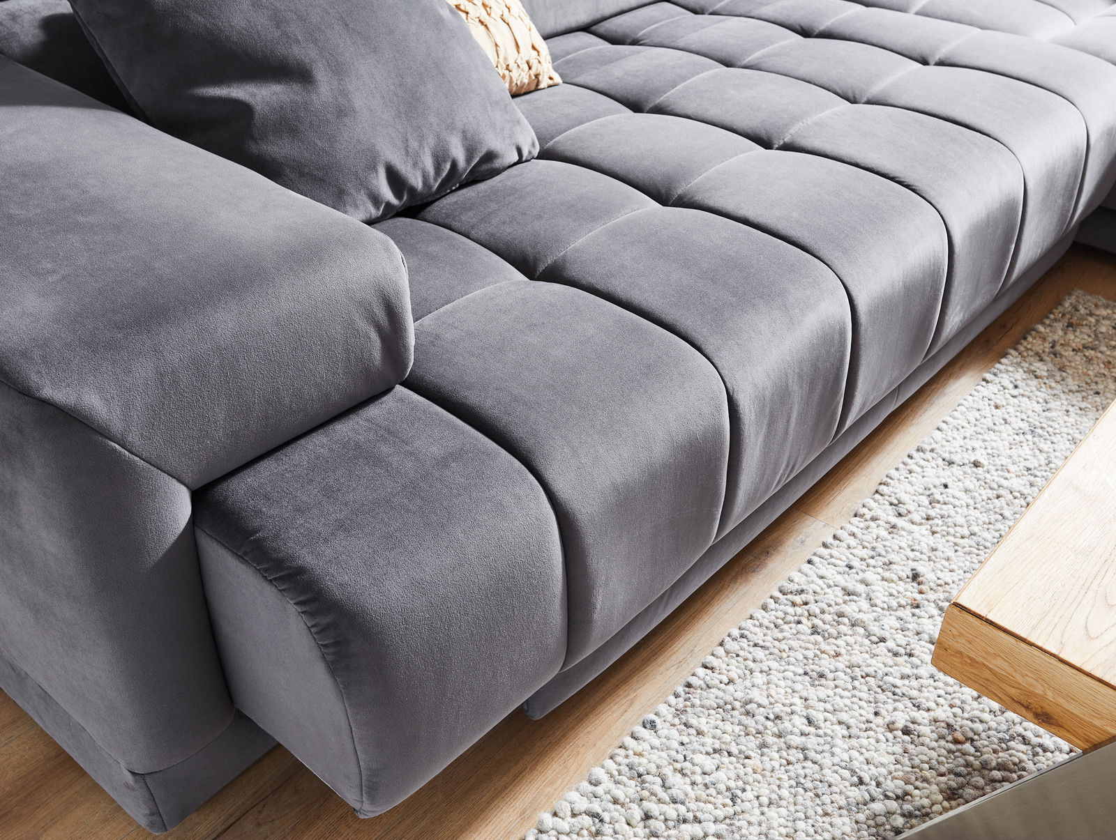 Graue Polsterrundecke aus Samt mit Kissen, Nahaufnahme von schräg oben, auf einem Holzboden und einem Teppich.