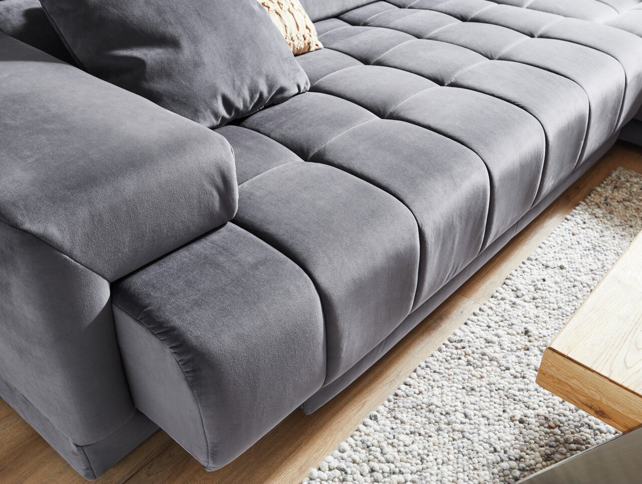 Graue Polsterrundecke aus Samt mit Kissen, Nahaufnahme von schräg oben, auf einem Holzboden und einem Teppich.