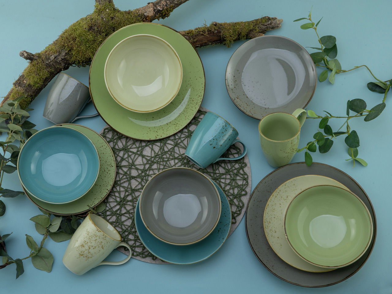 Vogelperspektive auf ein 16-teiliges Kombiservice-Set 'Nature' in verschiedenen Grüntönen, bestehend aus Tellern, Schalen und Tassen, dekoriert mit Moos und Zweigen.