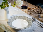 Modern gedeckter Esstisch mit gesprenkeltem Keramikteller, Besteck und Rotweinglas neben weißer Vase mit Blumen, in leicht schräger Seitenansicht von oben.