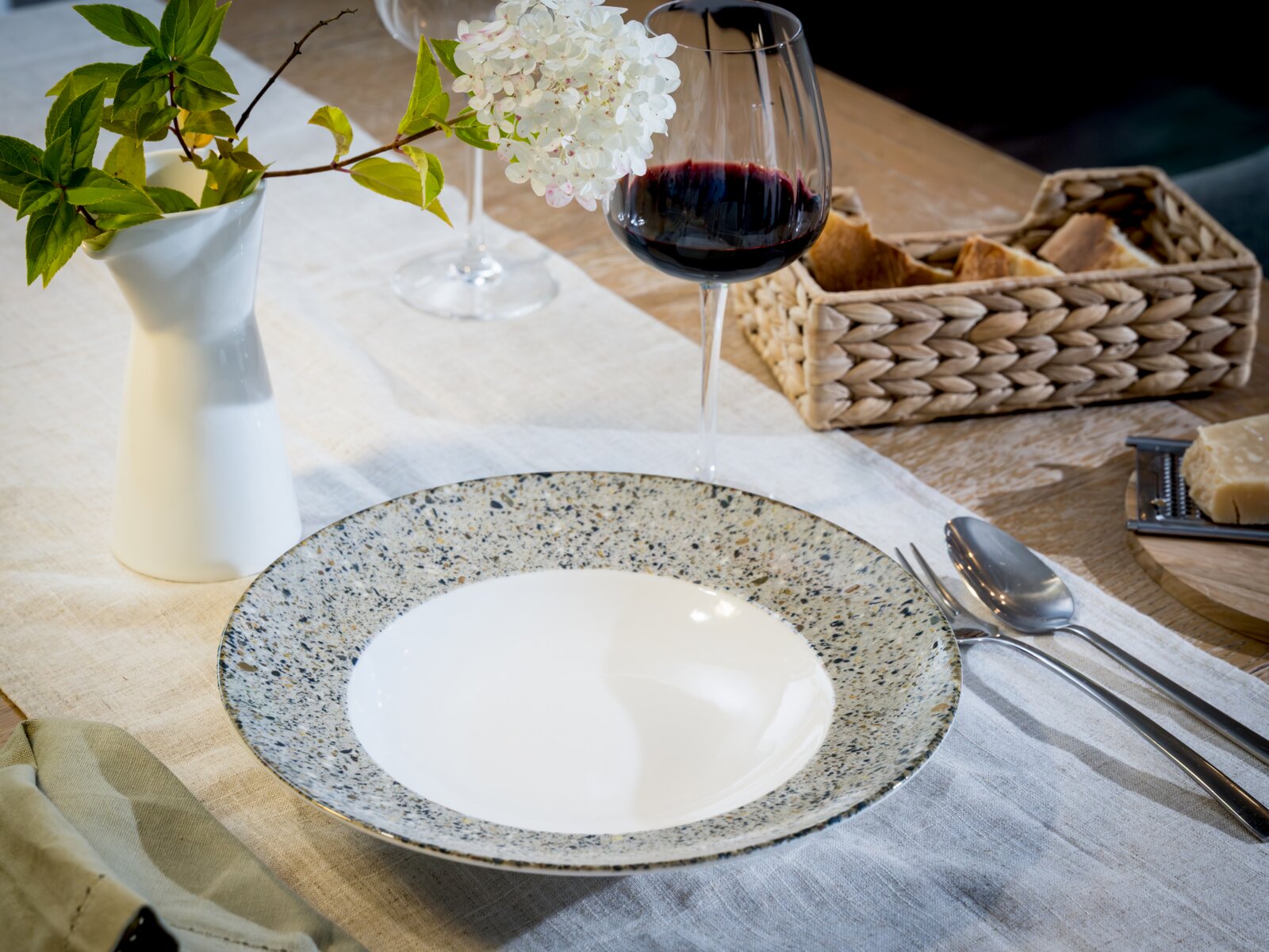 Modern gedeckter Esstisch mit gesprenkeltem Keramikteller, Besteck und Rotweinglas neben weißer Vase mit Blumen, in leicht schräger Seitenansicht von oben.