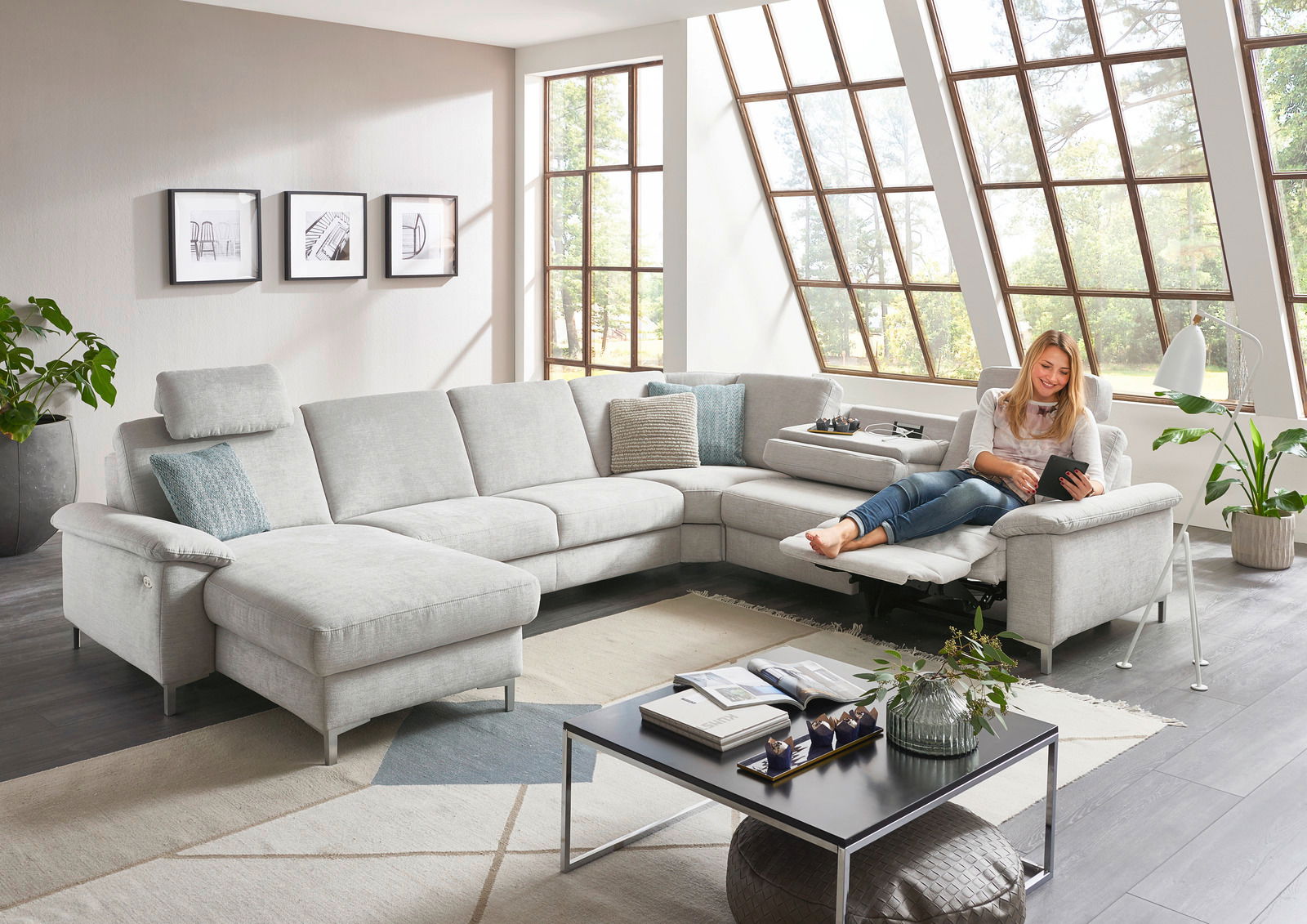 Helle Polsterrundecke in einem modernen Wohnzimmer mit großen Fenstern, von vorne betrachtet. Eine Person sitzt entspannt auf der Couch und liest ein Tablet.