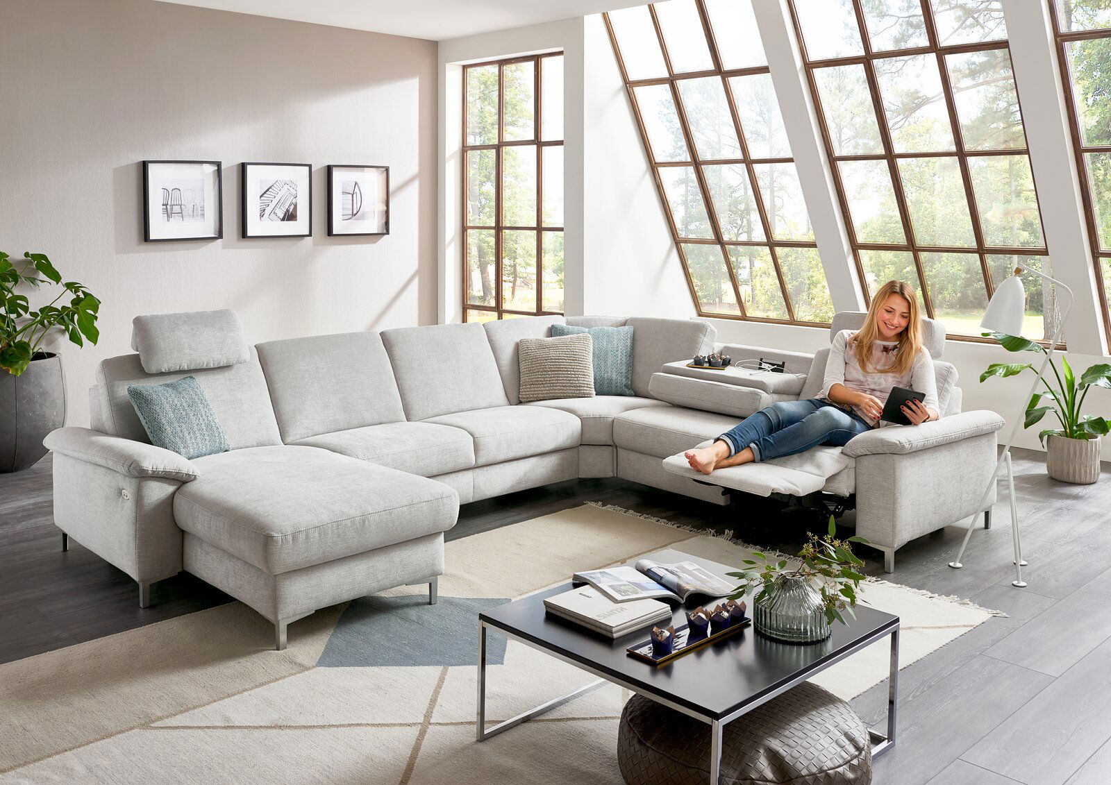 Helle Polsterrundecke in einem modernen Wohnzimmer mit großen Fenstern, von vorne betrachtet. Eine Person sitzt entspannt auf der Couch und liest ein Tablet.