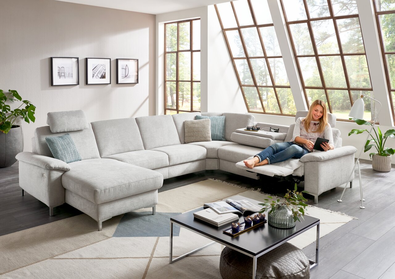 Helle Polsterrundecke in einem modernen Wohnzimmer mit großen Fenstern, von vorne betrachtet. Eine Person sitzt entspannt auf der Couch und liest ein Tablet.