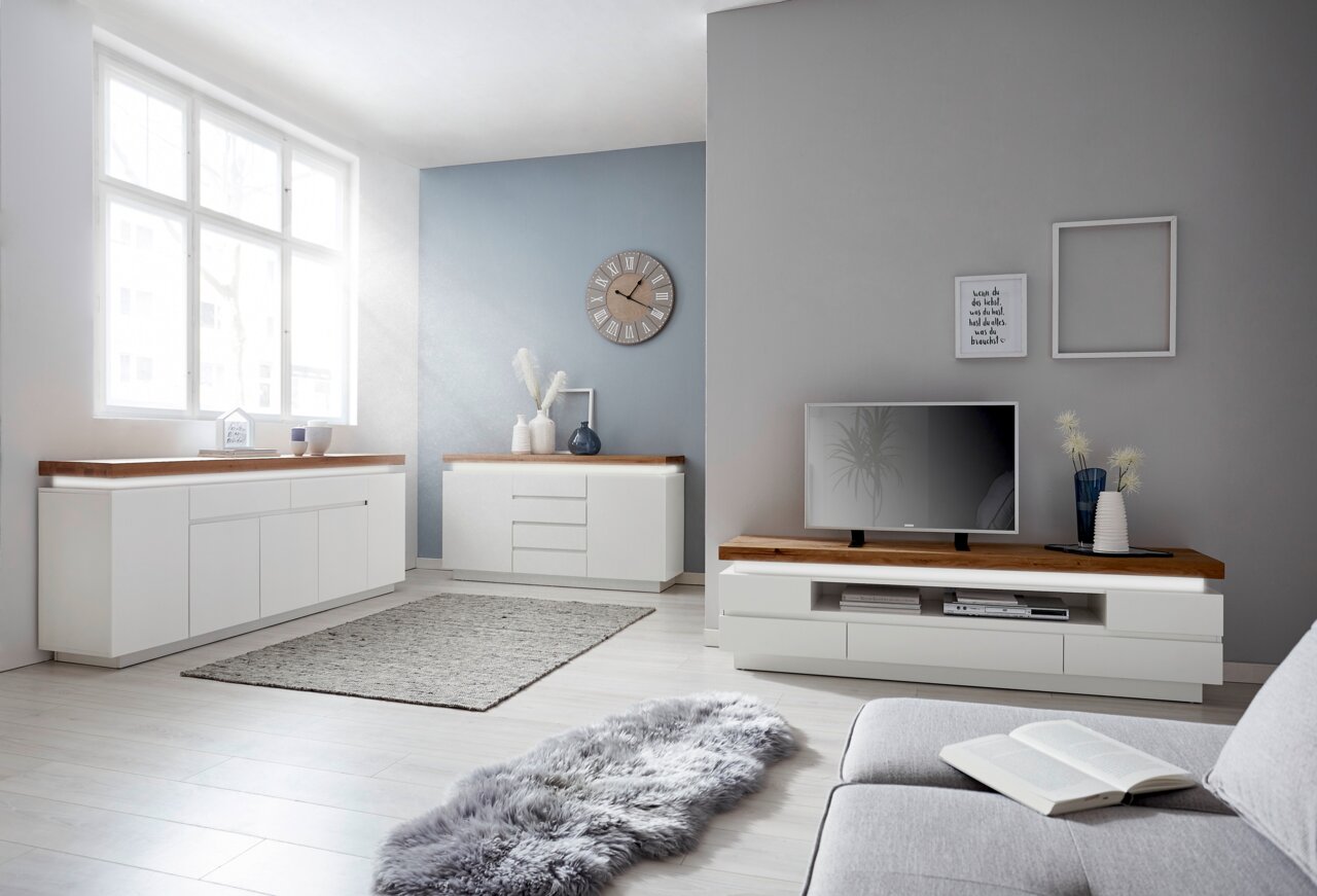 Novel XL-Sideboard ROMINA Modernes Wohnzimmer mit weißem TV-Lowboard, Sideboard und Kommode, alle mit Holzoberfläche. Perspektive von der Seite mit Blick auf die Möbel und ein großes Fenster, das viel Licht hereinlässt.