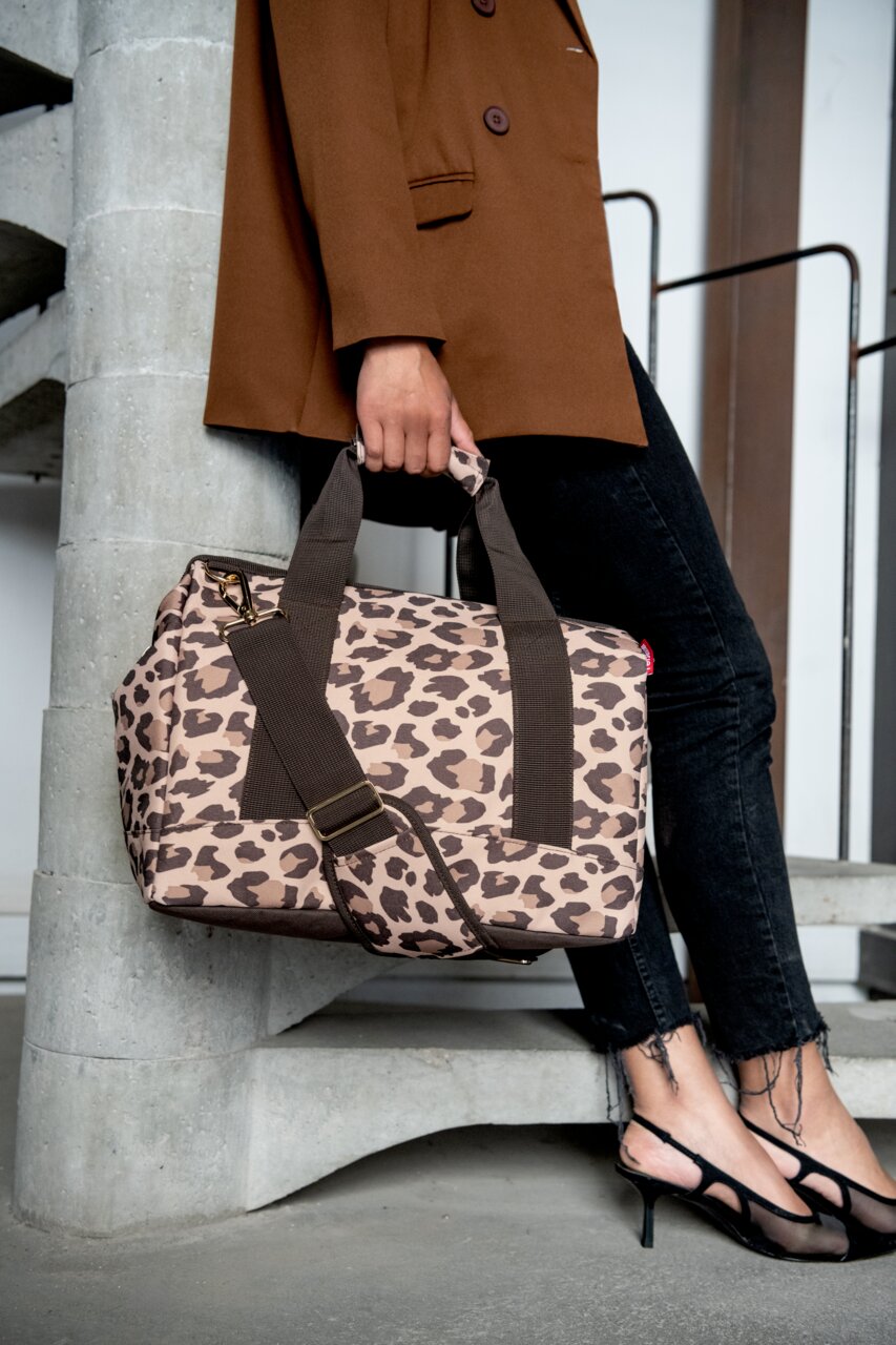 Reisenthel Allrounder M LEO MACCHIATO Leopardenmuster-Tasche in Macchiato-Farbe, getragen von einer Person in einem braunen Blazer und schwarzen Jeans, seitliche Perspektive.