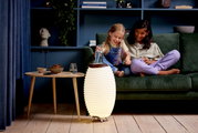 Akku-Leuchte und Lautsprecher in einem modernen Wohnzimmer, frontal betrachtet, neben einem Tisch und einem Sofa mit zwei lachenden Kindern.