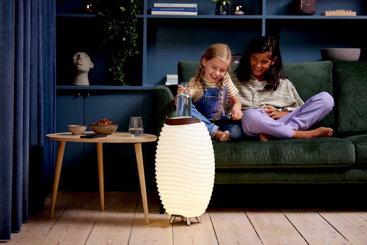 Akku-Leuchte und Lautsprecher in einem modernen Wohnzimmer, frontal betrachtet, neben einem Tisch und einem Sofa mit zwei lachenden Kindern.