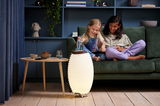 Akku-Leuchte und Lautsprecher in einem modernen Wohnzimmer, frontal betrachtet, neben einem Tisch und einem Sofa mit zwei lachenden Kindern.