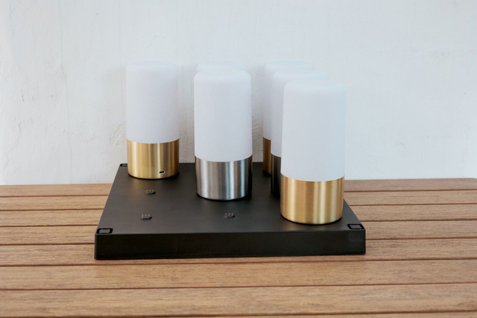 Newgarden Ladestation MAIA Moderne Tischleuchte mit vier zylindrischen, opalweißen Diffusoren auf schwarzer rechteckiger Basis, Metallfüße in Gold und Silber, fotografiert in frontaler leicht erhöhter Perspektive auf Holztisch vor weißer Wand