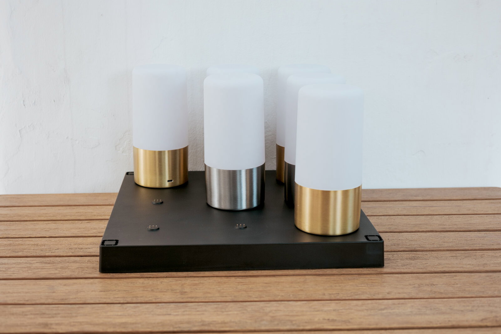 Newgarden Ladestation MAIA Moderne Tischleuchte mit vier zylindrischen, opalweißen Diffusoren auf schwarzer rechteckiger Basis, Metallfüße in Gold und Silber, fotografiert in frontaler leicht erhöhter Perspektive auf Holztisch vor weißer Wand