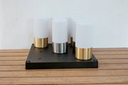 Moderne Tischleuchte mit vier zylindrischen, opalweißen Diffusoren auf schwarzer rechteckiger Basis, Metallfüße in Gold und Silber, fotografiert in frontaler leicht erhöhter Perspektive auf Holztisch vor weißer Wand