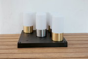 Moderne Tischleuchte mit vier zylindrischen, opalweißen Diffusoren auf schwarzer rechteckiger Basis, Metallfüße in Gold und Silber, fotografiert in frontaler leicht erhöhter Perspektive auf Holztisch vor weißer Wand