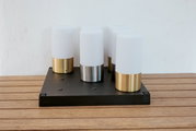 Moderne tafellamp met vier cilindervormige, opaalwitte lichtverspreiders op een zwarte rechthoekige voet, metalen poten in goud en zilver, gefotografeerd vanuit een licht verhoogd vooraanzicht op een houten tafel voor een witte muur
