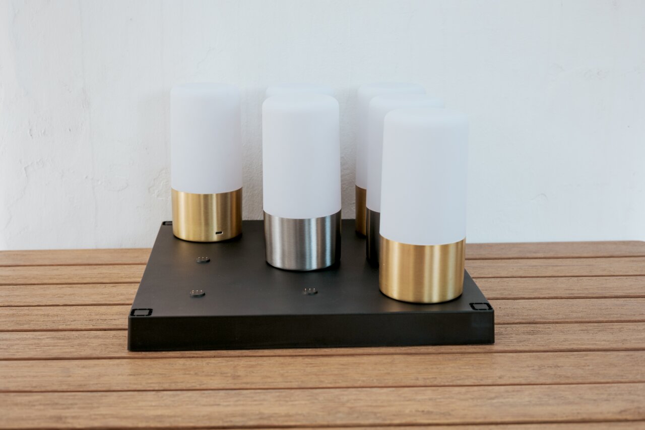Moderne tafellamp met vier cilindervormige, opaalwitte lichtverspreiders op een zwarte rechthoekige voet, metalen poten in goud en zilver, gefotografeerd vanuit een licht verhoogd vooraanzicht op een houten tafel voor een witte muur