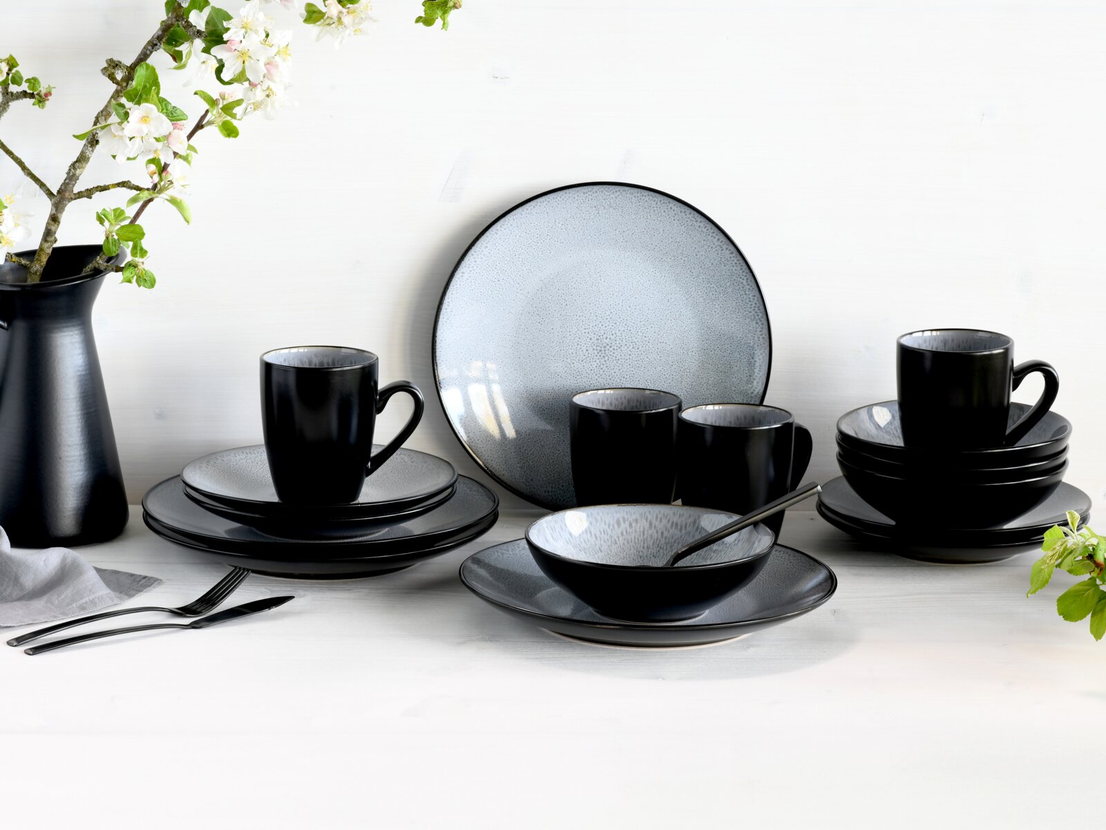 Elegantes 16-teiliges Kombiservice in Schwarz und Grau, bestehend aus Tellern, Tassen und Schalen, auf einem Tisch mit Blumenarrangement, aus frontaler Perspektive.