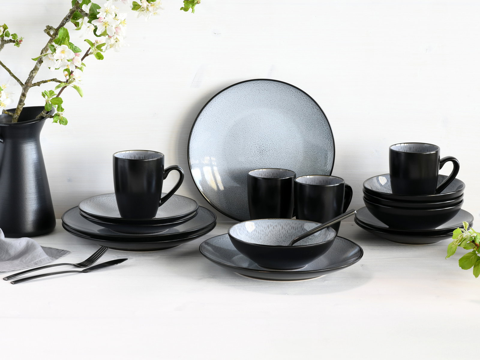 Creatable Kombiservice 16-tlg MALMÖ Elegantes 16-teiliges Kombiservice in Schwarz und Grau, bestehend aus Tellern, Tassen und Schalen, auf einem Tisch mit Blumenarrangement, aus frontaler Perspektive.