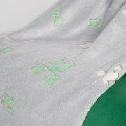 Close-up vanuit een schuin perspectief: zachte grijze fleecedeken met groene pixel-/creeper-motieven en een wit koord langs de rand op een groene achtergrond
