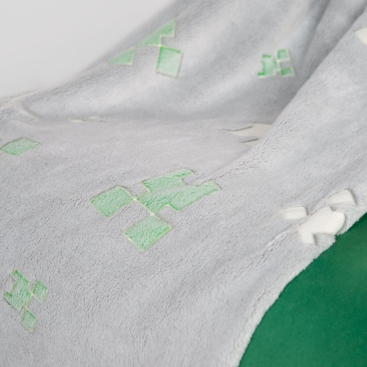 Close-up vanuit een schuin perspectief: zachte grijze fleecedeken met groene pixel-/creeper-motieven en een wit koord langs de rand op een groene achtergrond