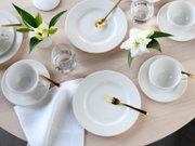 Modern gedeckter Tisch mit weißem Porzellan-Geschirr, Tassen und Tellern, goldfarbenem Besteck, Wassergläsern und weißen Lilien auf hellem Holztisch, fotografiert aus der Vogelperspektive (Draufsicht).