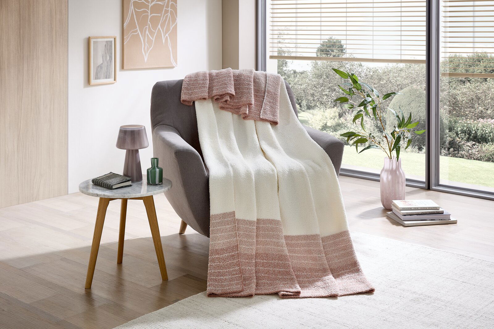 Linneweber Wohndecke SAMMY 150x200cm Gemütliche Wohndecke in Creme und Rosa auf einem grauen Sessel, mit Blick auf ein modernes Wohnzimmer mit großen Fenstern.