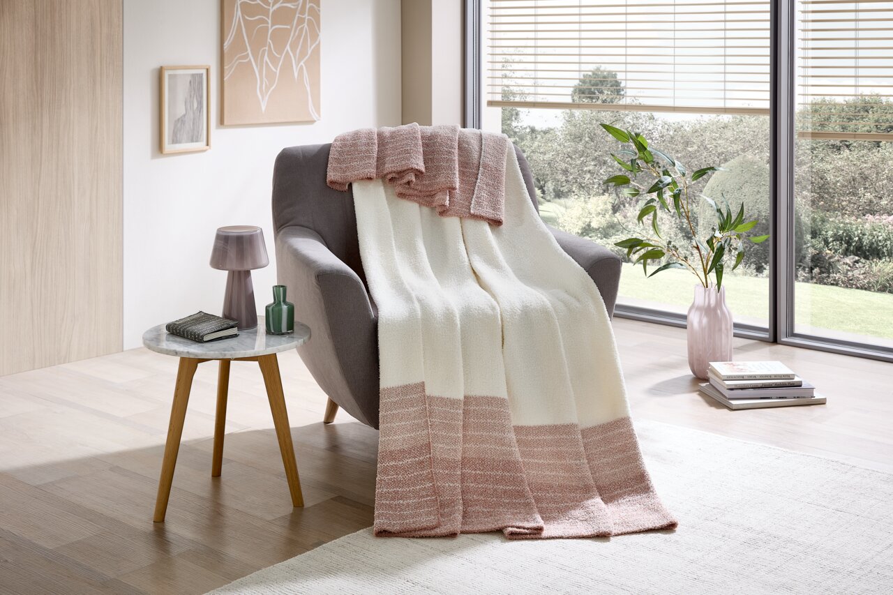 Gemütliche Wohndecke in Creme und Rosa auf einem grauen Sessel, mit Blick auf ein modernes Wohnzimmer mit großen Fenstern.