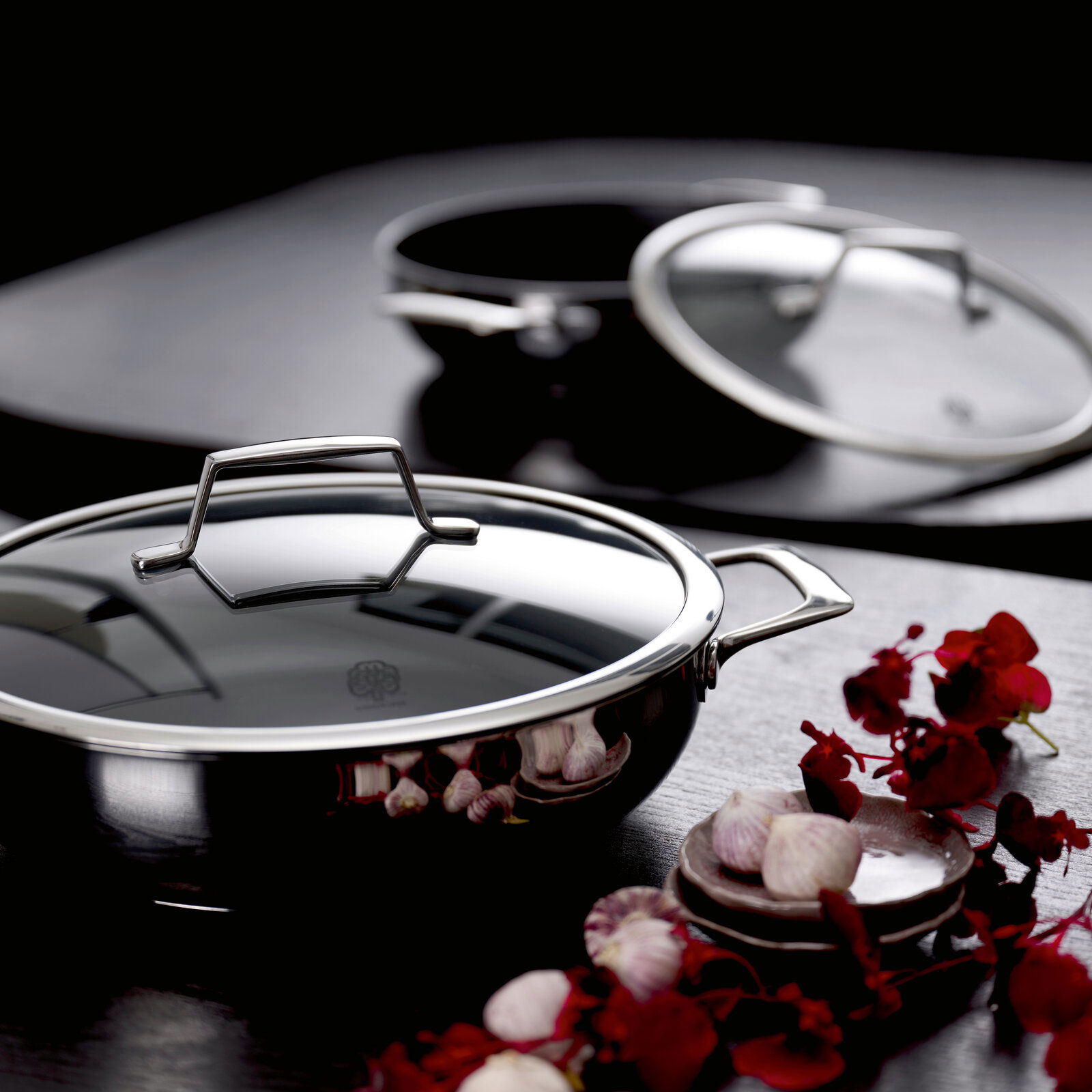 Glänzender Edelstahl-Kochtopf mit Glasdeckel und Metallgriffen auf dunkler Oberfläche, im Vordergrund aus leicht erhöhter Frontansicht; im Hintergrund weiterer Topf, daneben Knoblauch und rote Blüten.