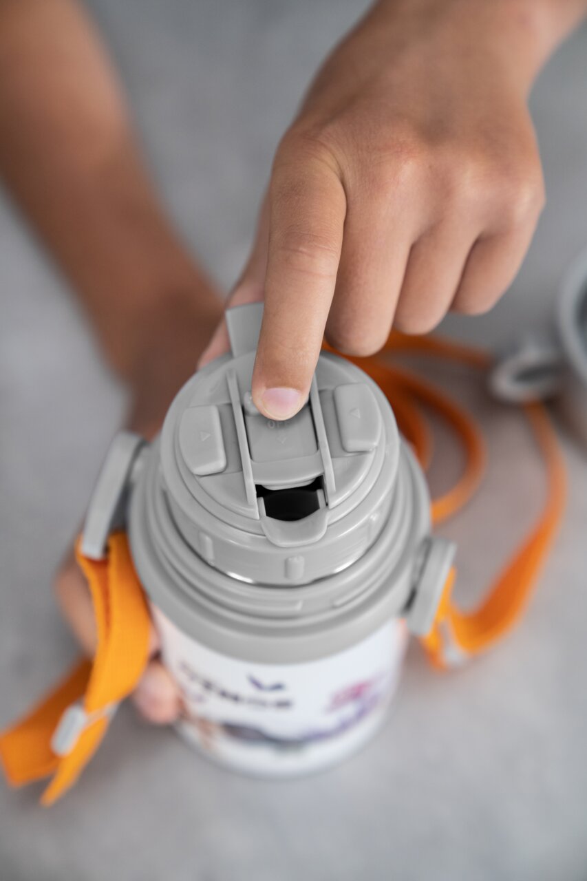 Nahaufnahme einer grauen Thermoflasche mit Becher und orangem Tragegurt, von oben betrachtet, mit einer Hand, die den Verschluss öffnet.